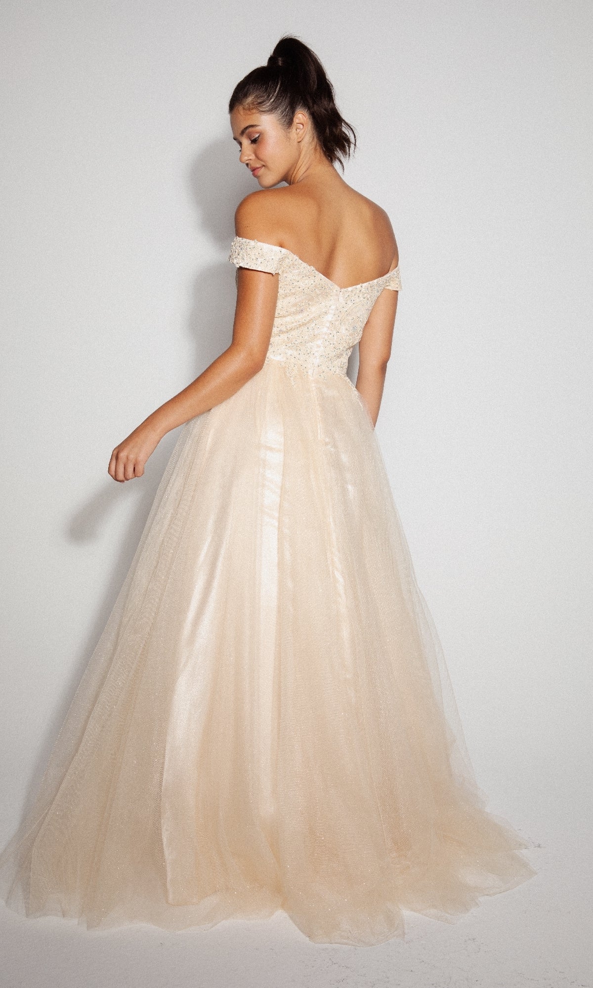 A woman in the Off-Shoulder Glitter Prom Ball Gown: Eureka 9899, featuring a light beige tulle skirt, stands with her back turned and gazes over her shoulder.